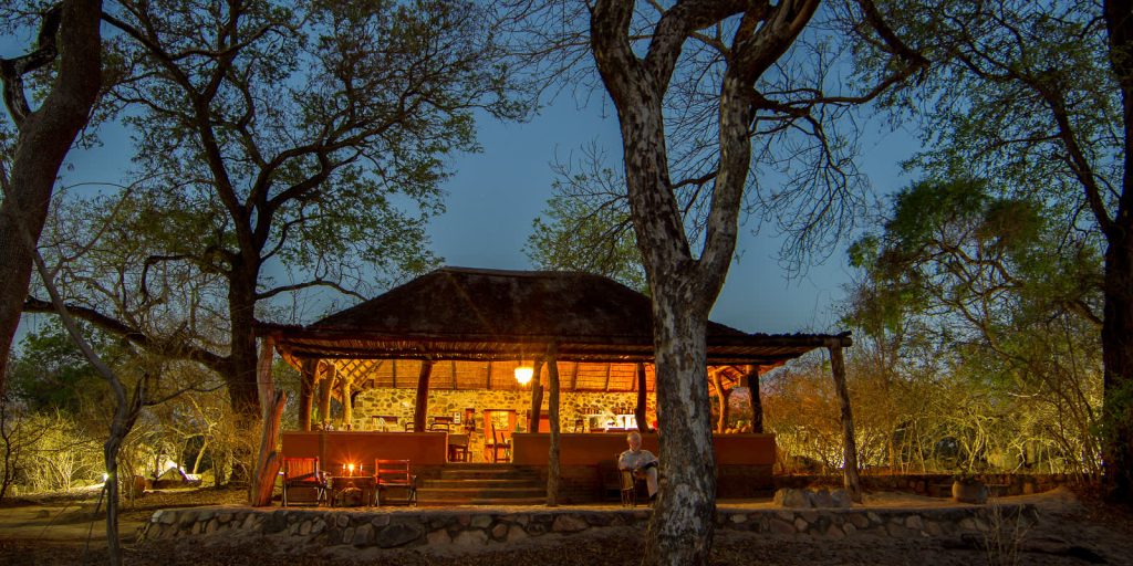 Photo of a luxury tented camp with thatched roofs and wooden decks within the borders of the Majete Wildlife Reserve. Lush green vegetation surrounds the camp, and mountains are visible in the distance.