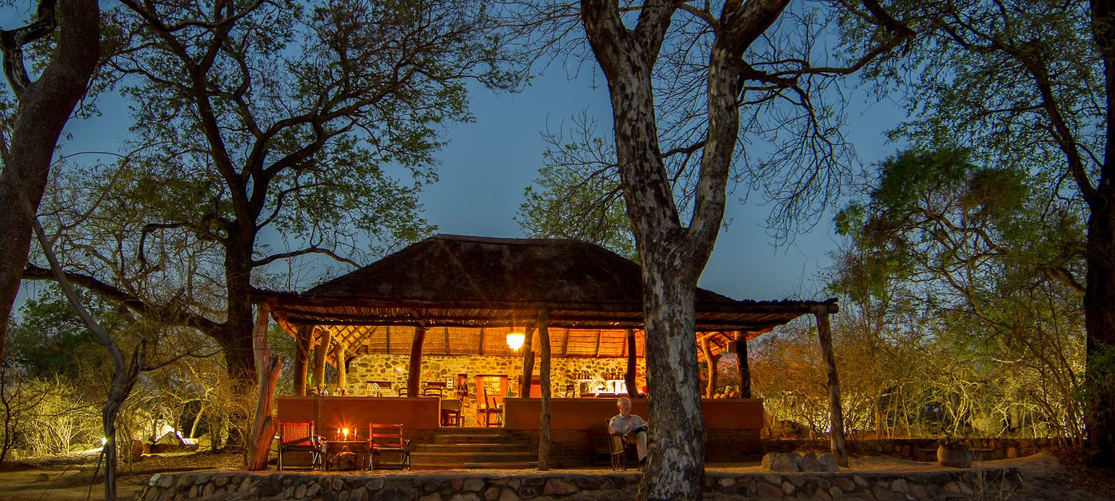 Photo of a luxury tented camp with thatched roofs and wooden decks within the borders of the Majete Wildlife Reserve. Lush green vegetation surrounds the camp, and mountains are visible in the distance.